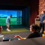 Man in blue shirt holding club on Full Swing Golf Simulator with group of friends watching him